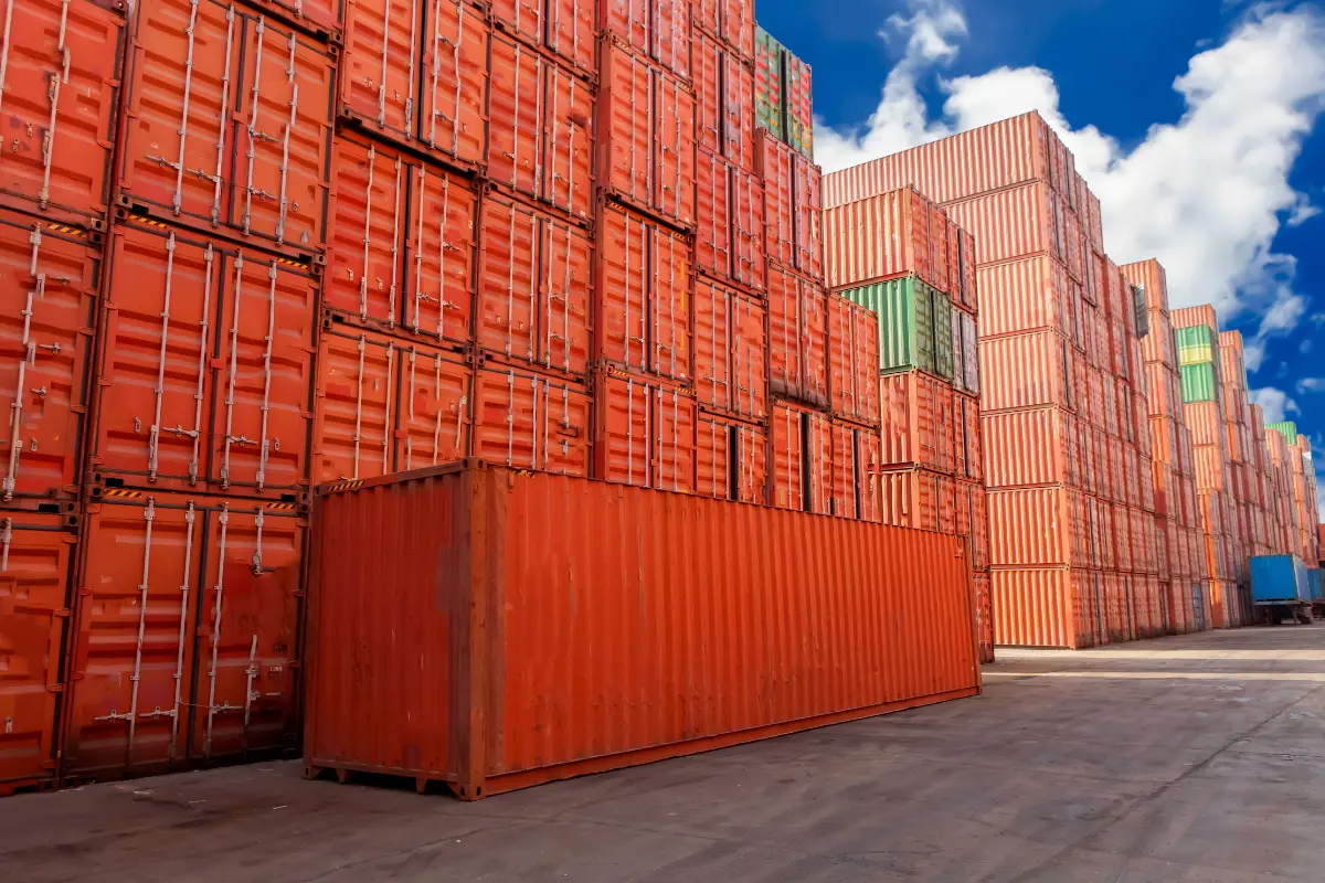 Stacked 40-foot cargo containers in bright orange and various colors at a shipping yard under a blue sky.